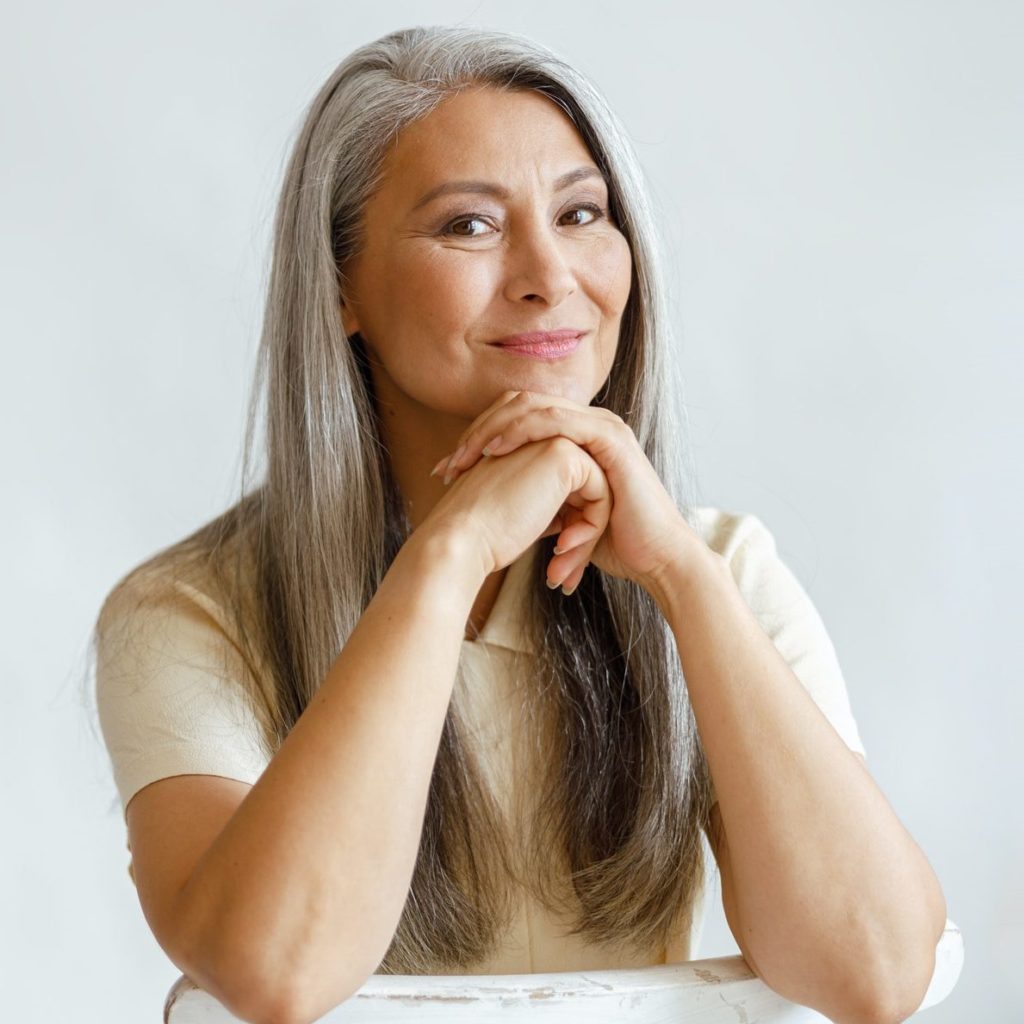Happy middle aged woman with silver hair sits leaning on hands on light background